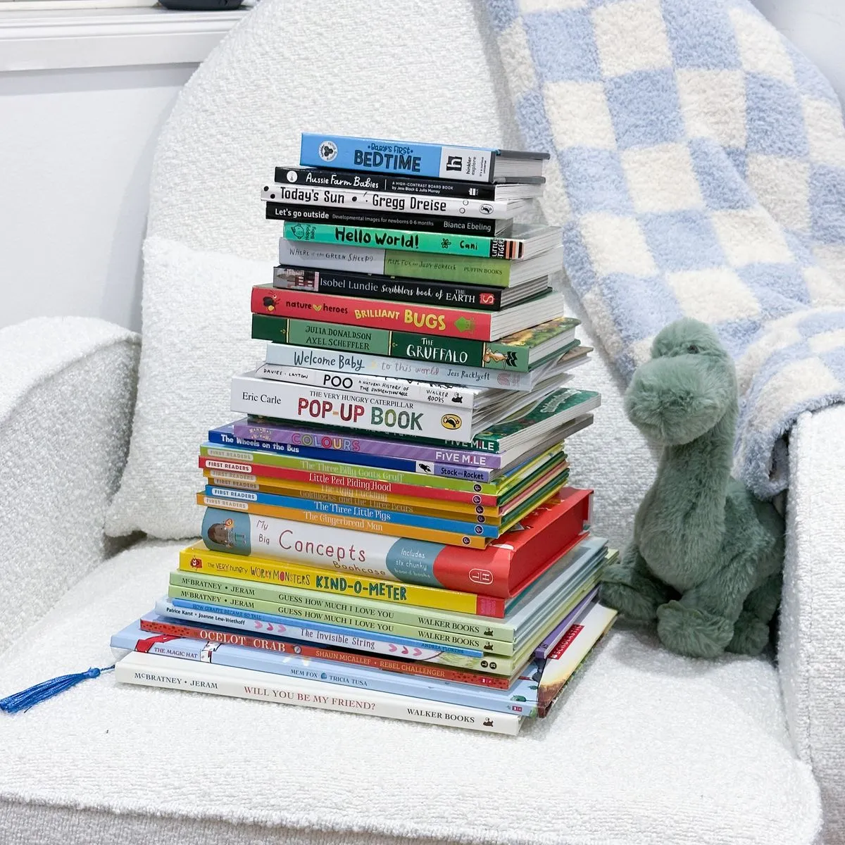 A tall stack of children’s books on a chair, beside a toy dinosaur.
